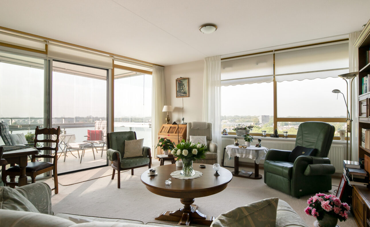 Woonkamer met grote ramen, comfortabele stoelen, een ronde houten tafel met bloemen en uitzicht op een balkon met zitje.