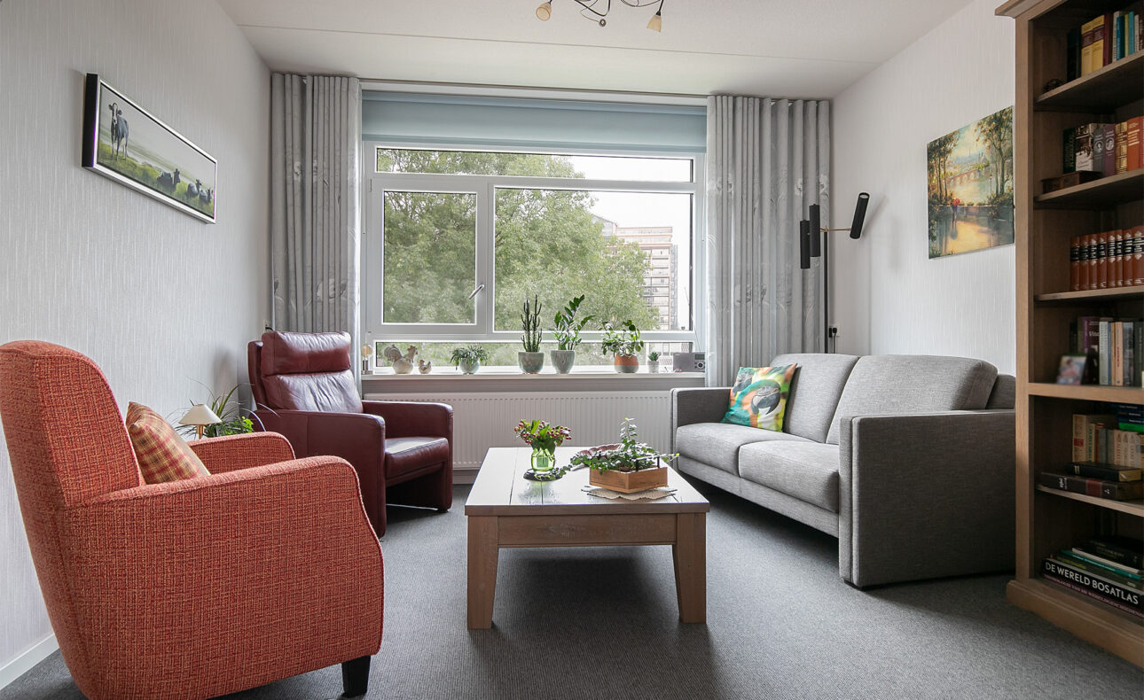 Woonkamer met een grijze bank, rode en bruine fauteuils, houten salontafel, boekenplank en grote ramen met uitzicht op bomen.