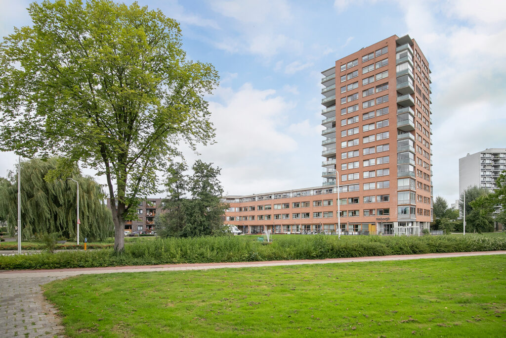 Hoog woongebouw met rode bakstenen en balkons, omgeven door groen gras en bomen onder een bewolkte hemel.