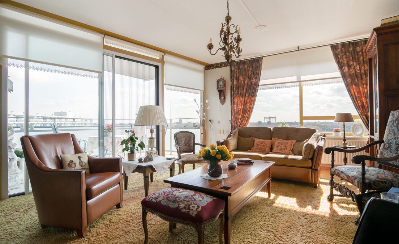 Woonkamer met grote ramen, een bruine bank, leren fauteuil, houten salontafel en een kroonluchter aan het plafond.