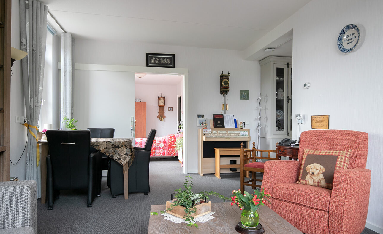 Woonkamer met rode fauteuil, houten tafel met bloemen, zwarte stoelen en een kast. Deur leidt naar een andere kamer.
