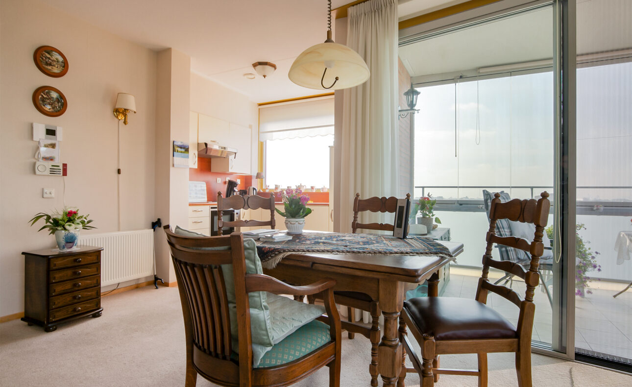 Eetkamer met houten tafel en stoelen, bloemen op tafel, grote ramen met uitzicht op balkon en veel natuurlijk licht.