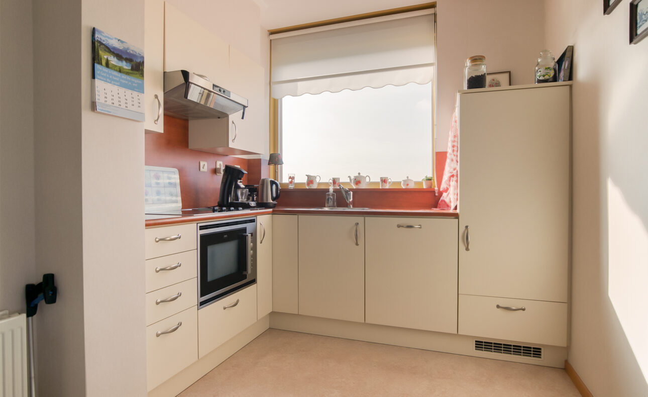 Kleine, lichte keuken met witte kasten, een fornuis, oven en gootsteen. Er is een groot raam met planten op de vensterbank.