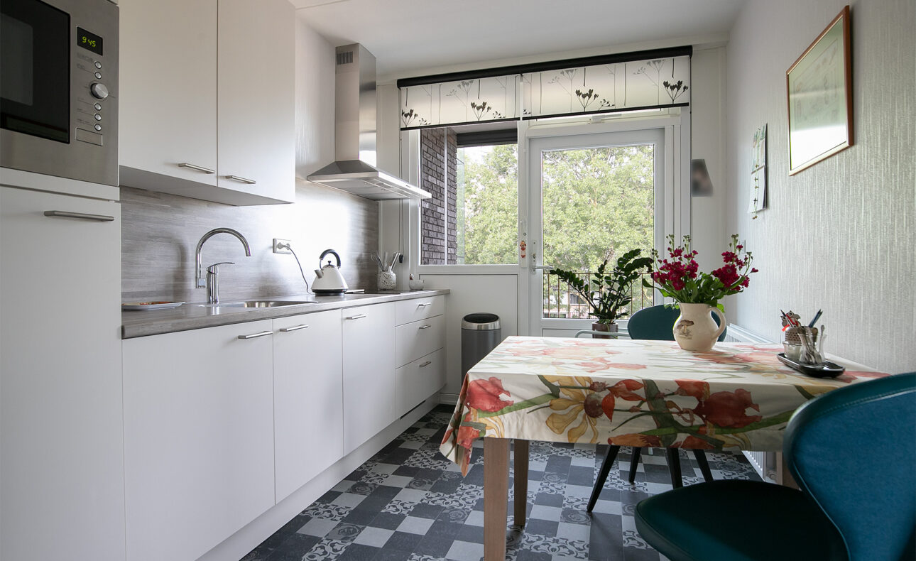 Keuken met witte kasten, een tafel met bloemenkleed, twee stoelen en een raam met uitzicht op groene bomen.