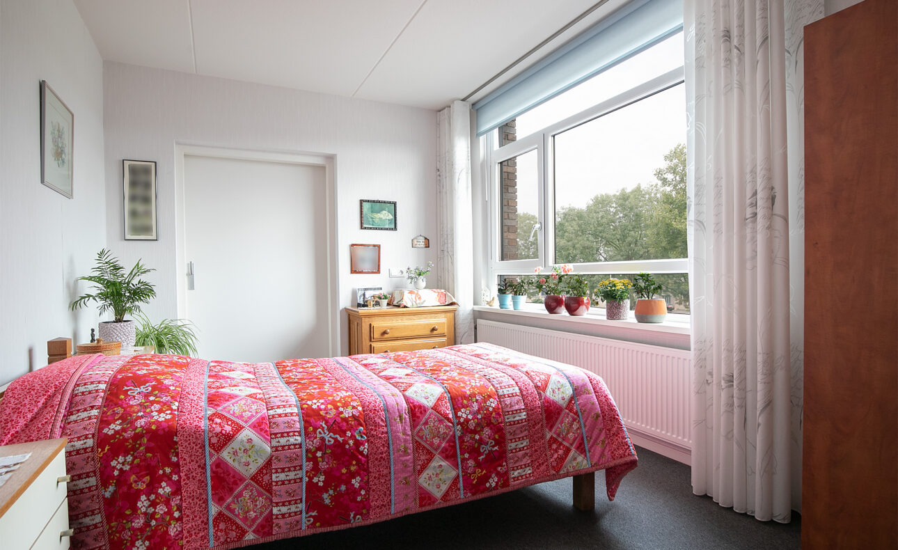Slaapkamer met een kleurrijk rood dekbed, houten meubels, planten op de vensterbank en een groot raam met uitzicht op groen.