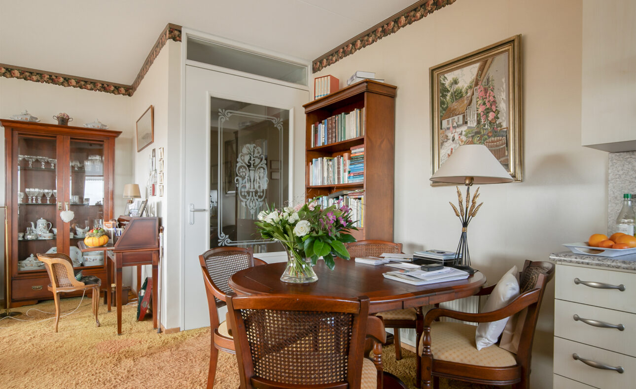 Een gezellige woonkamer met een ronde houten tafel, omringd door stoelen. Er staat een boekenkast en een kast met servies.