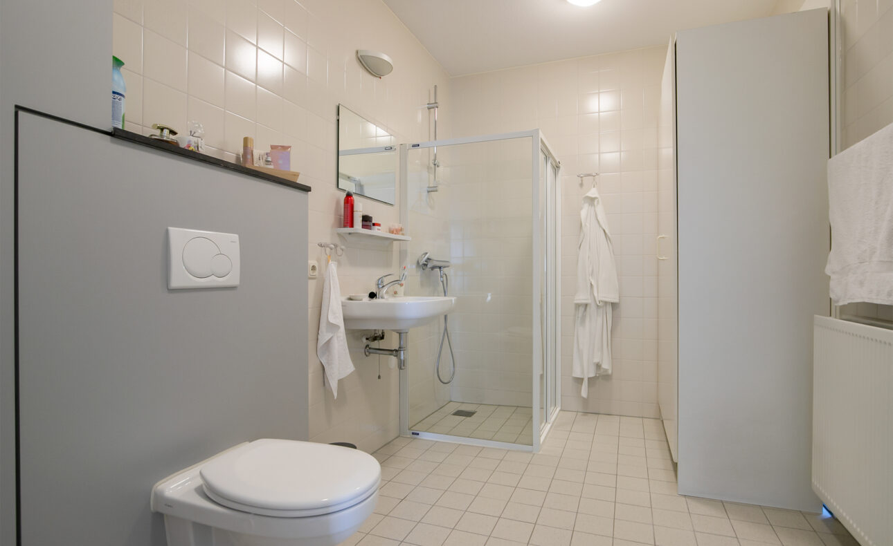 Een moderne badkamer met een toilet, wastafel en glazen douchecabine. De muren en vloer zijn betegeld in lichte kleuren.