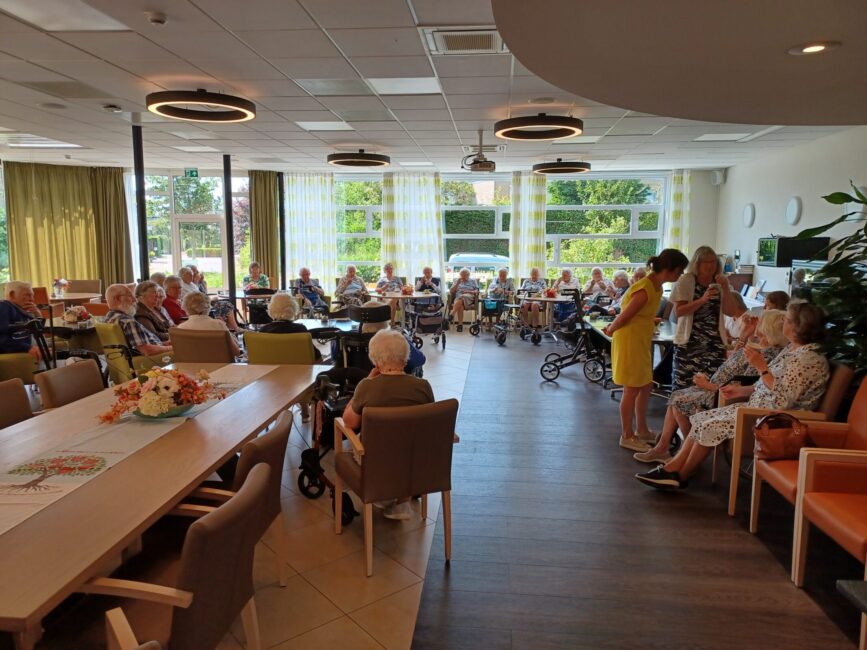 Groep ouderen zit in een lichte gemeenschappelijke ruimte met tafels en stoelen. Twee vrouwen staan en praten met elkaar.