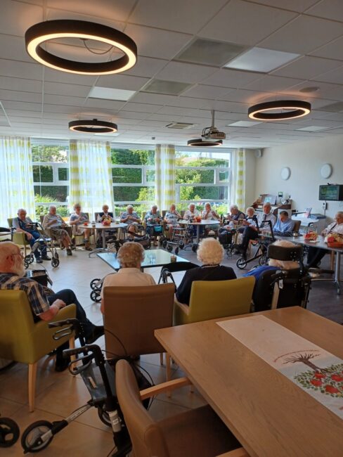 Groep ouderen zit in een gemeenschappelijke ruimte van een verzorgingstehuis, omringd door tafels en rollators.