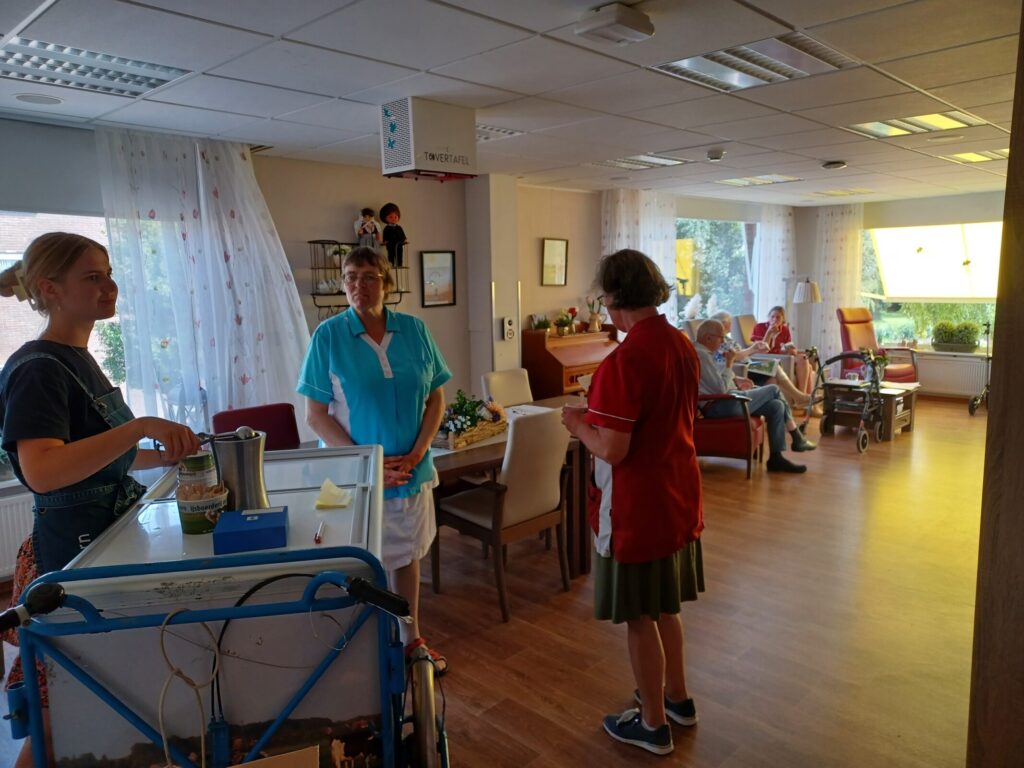Een zorginstelling met personeel en bewoners in een gemeenschappelijke ruimte. Mensen praten en zitten in stoelen.