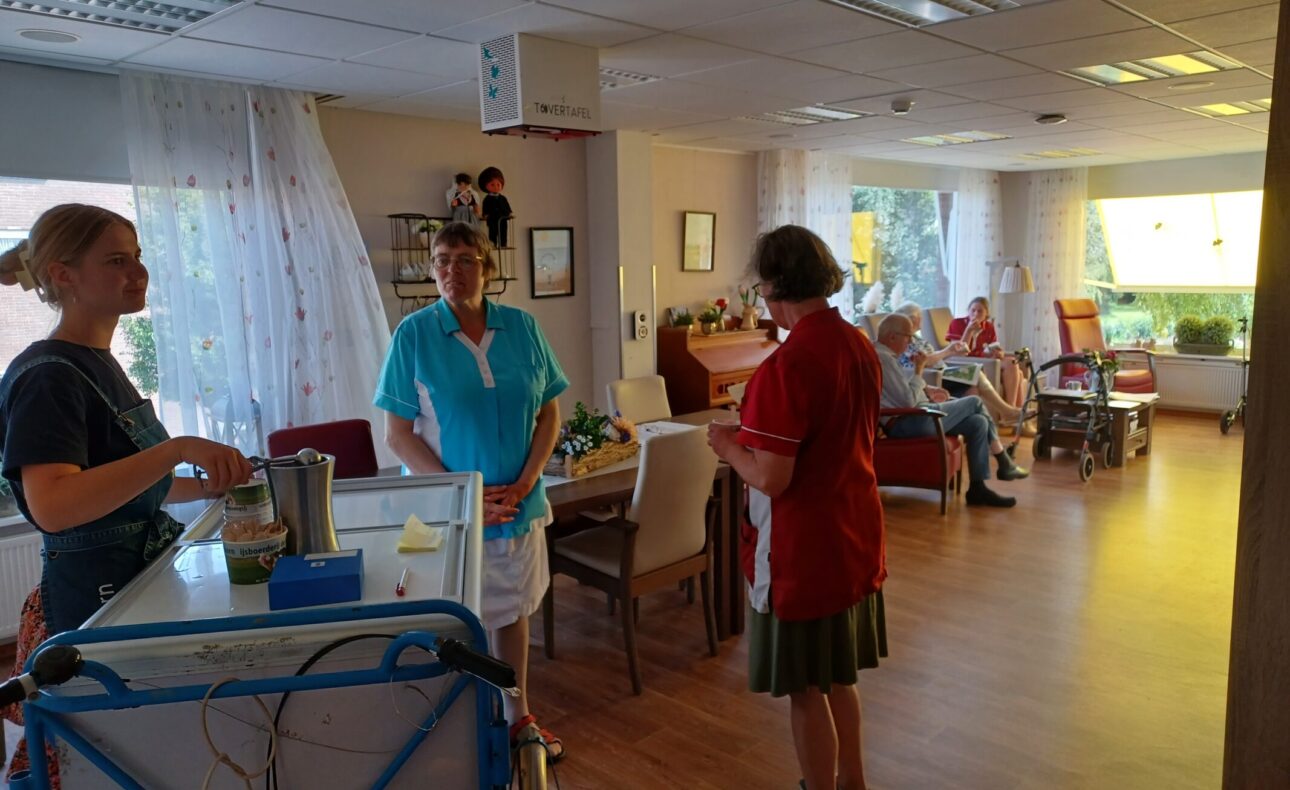 Een zorginstelling met personeel en bewoners in een gemeenschappelijke ruimte. Mensen praten en zitten in stoelen.