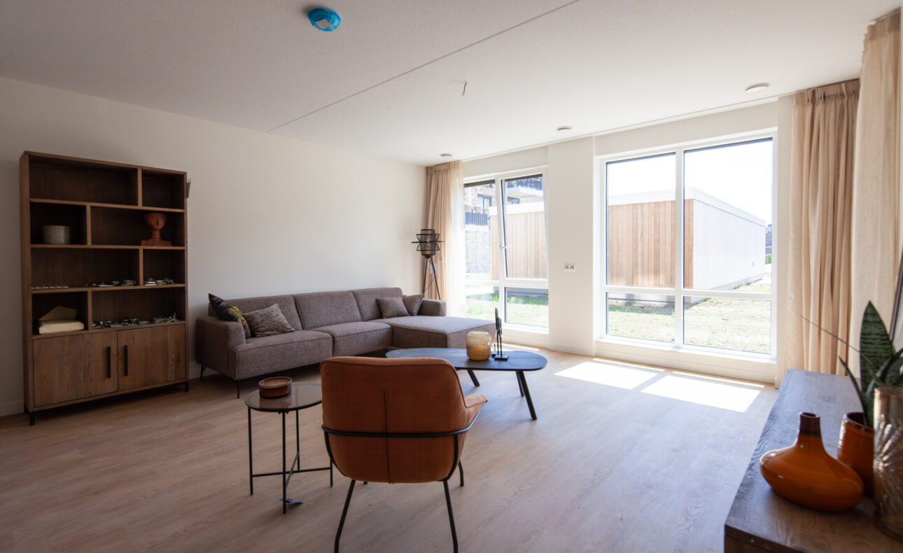 Op de foto is een woonkamer te zien van één van de appartementen. De woning heeft een grote raampartij waardoor er veel licht naar binnen valt. In de hoek van de woning staat een grijze hoekbank, daar tegenover staat een leren fauteuil.