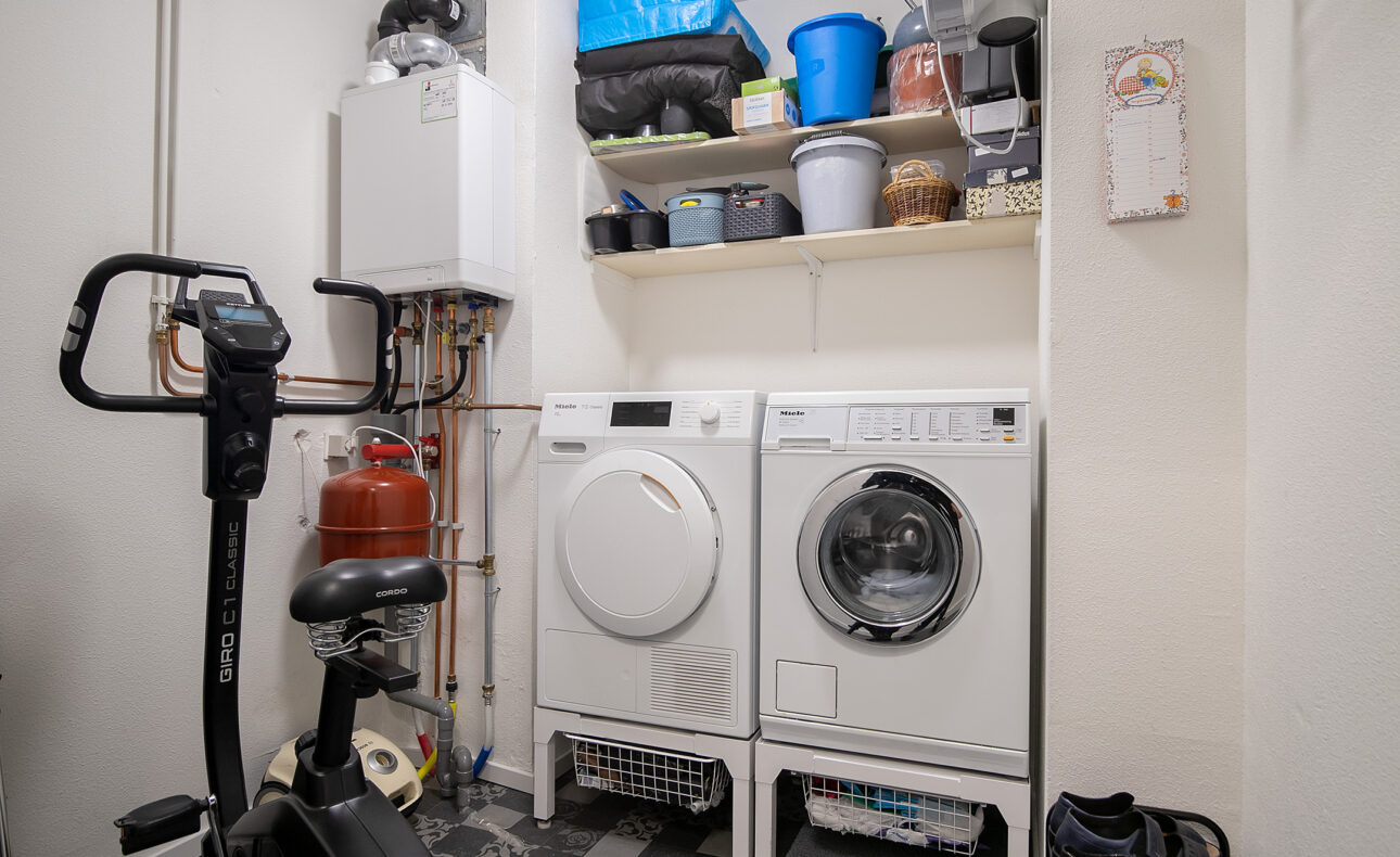 Waskamer met een wasmachine, droger, planken met schoonmaakmiddelen en een hometrainer op een zwart-wit geblokte vloer.