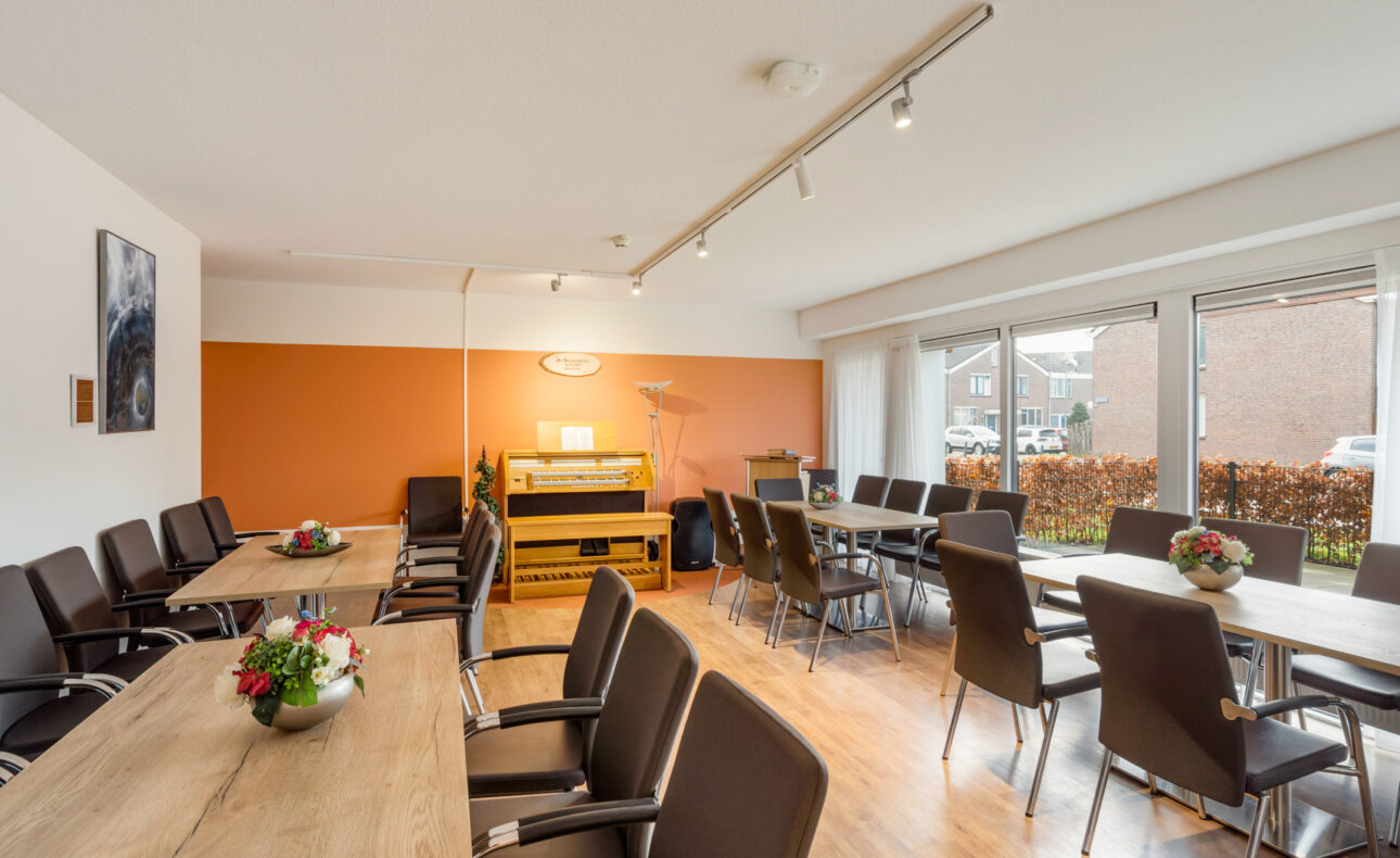 Vergaderruimte met houten tafels en zwarte stoelen, oranje accentmuur, en grote ramen die uitzicht bieden op een tuin.