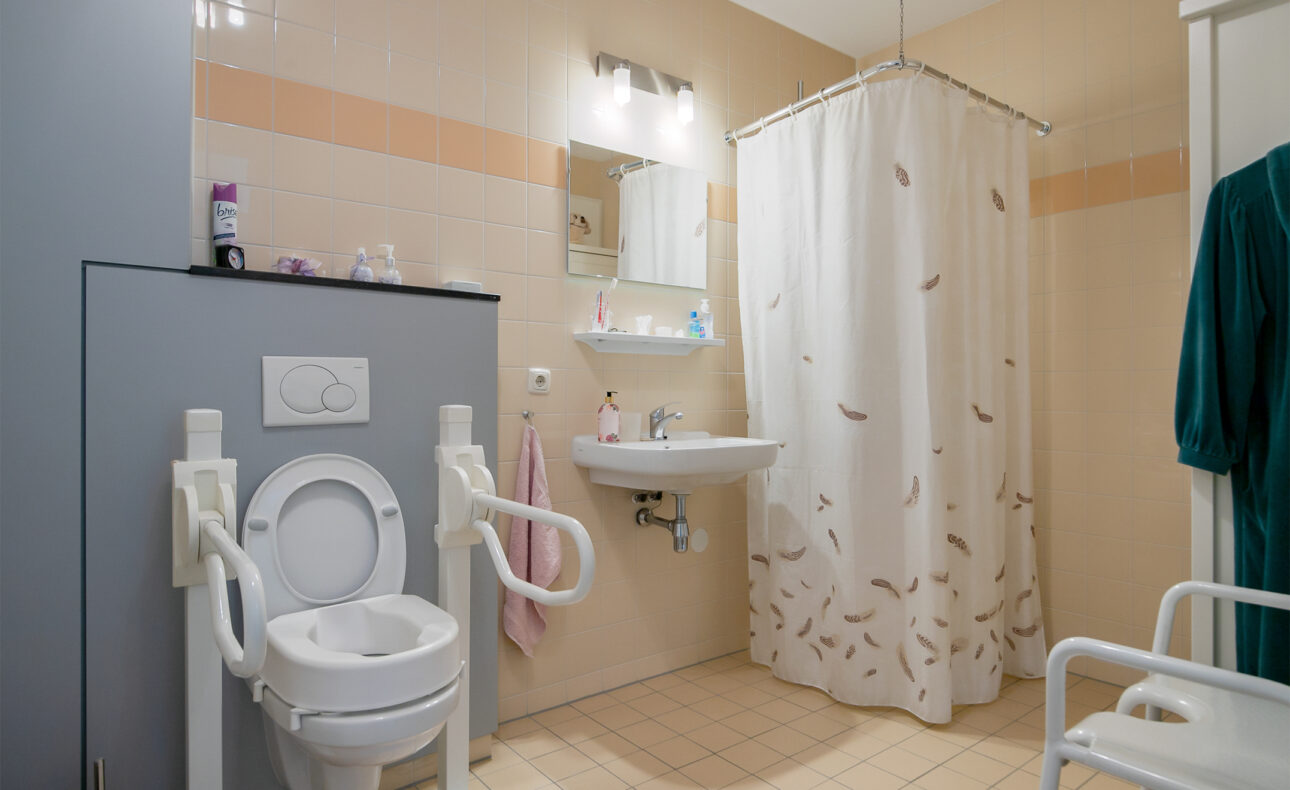 Badkamer met toilet met armsteunen, wastafel met spiegel, en douche met gordijn. Beige tegels en diverse toiletartikelen aanwezig.
