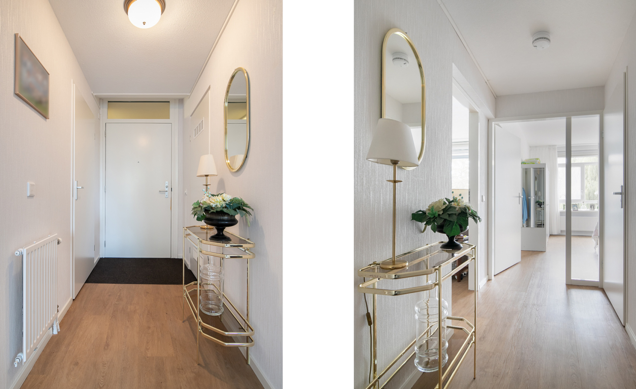 Een lichte gang met houten vloer, een ronde spiegel, een glazen tafel met bloemen en een lamp, leidend naar een open deur.