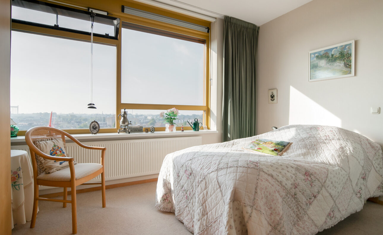 Een lichte slaapkamer met een groot raam, een bed met een gebloemd dekbedovertrek, een stoel en enkele planten op de vensterbank.
