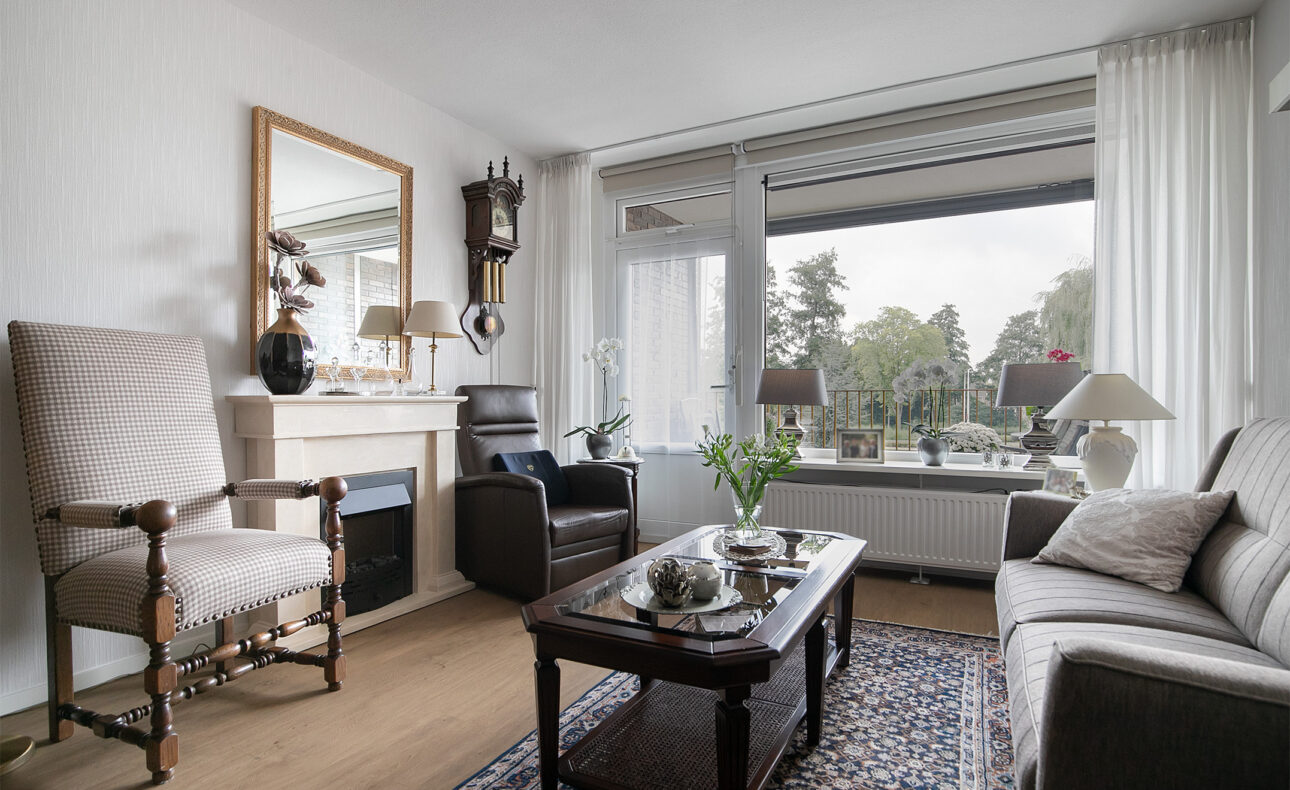 Woonkamer met klassieke meubels, een groot raam met uitzicht op bomen, en een tapijt op de houten vloer.