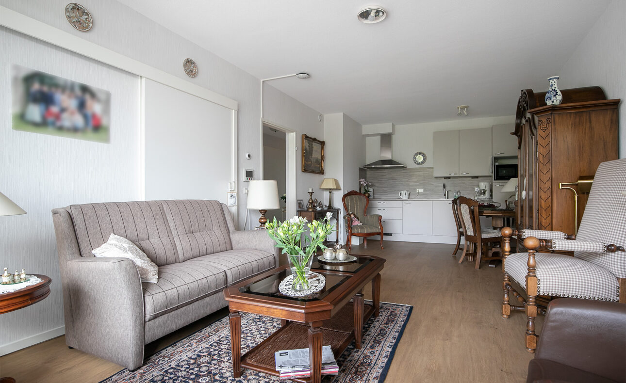Woonkamer met een bank, fauteuils, houten salontafel, bloemenvaas, tapijt en open keuken met witte kasten en apparatuur.