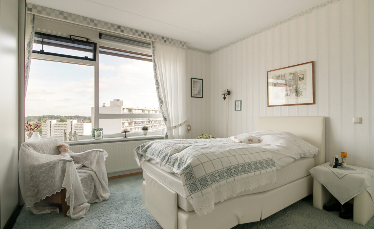 Slaapkamer met een groot raam, een bed met wit beddengoed, een stoel met een deken en een schilderij aan de muur.