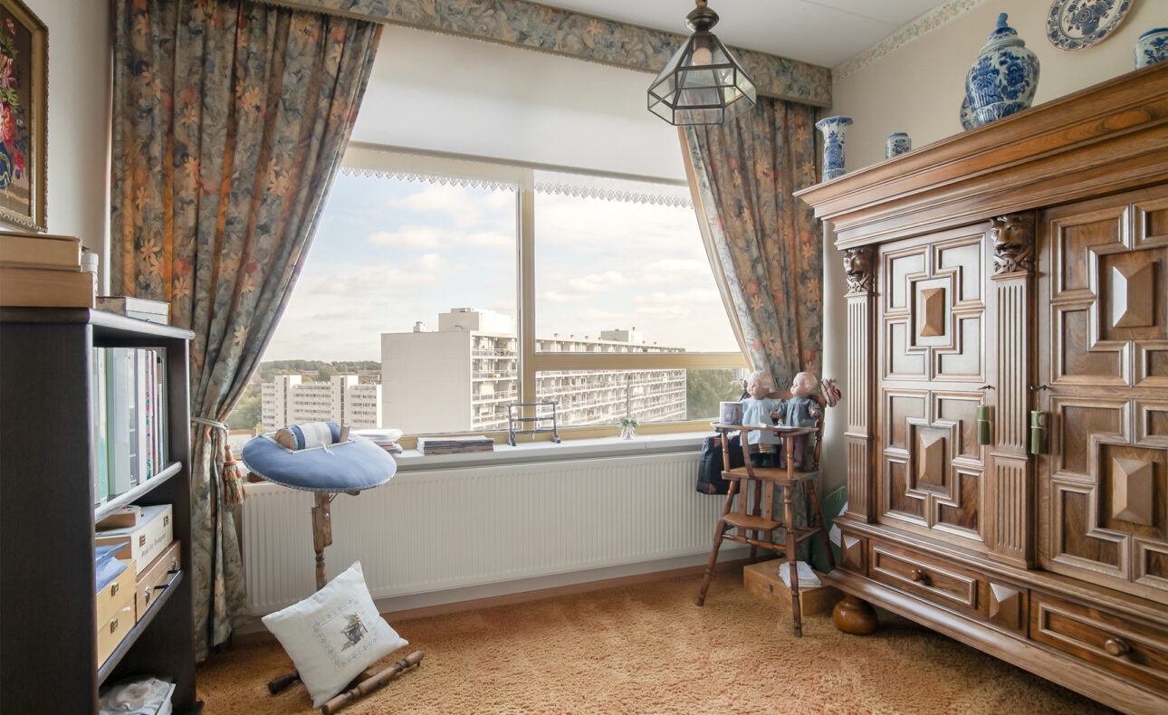 Een gezellige kamer met een grote houten kast, een raam met uitzicht op gebouwen, en een stoel bij een klein tafeltje.