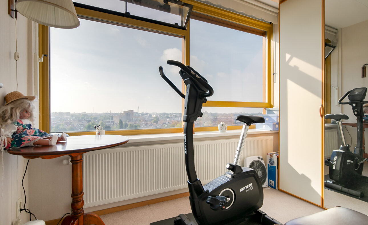 Een hometrainer staat in een kamer met een groot raam dat uitzicht biedt op een stad. Er is een tafel met een pop erop.
