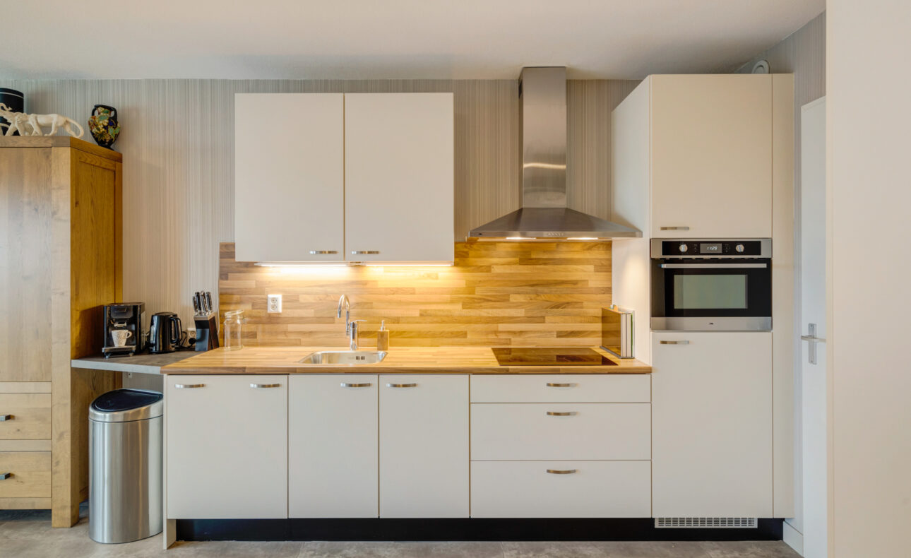 Moderne keuken met witte kasten, houten werkblad, ingebouwde oven en afzuigkap. Links een koffiezetapparaat en prullenbak.