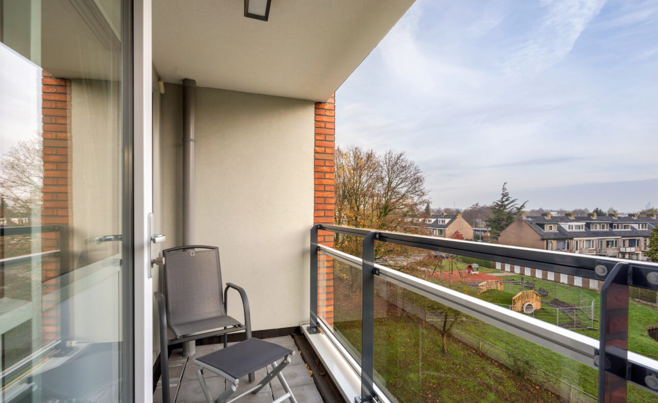 Balkon van een modern appartement met twee stoelen, uitzicht op een groene tuin en een woonwijk in de verte onder een blauwe lucht.