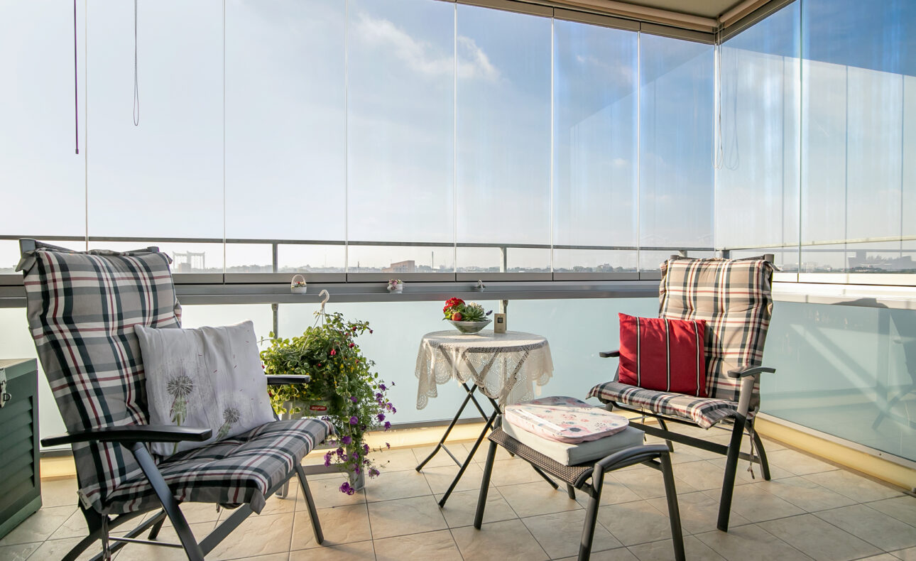 Twee comfortabele stoelen met kussens op een balkon, naast een klein tafeltje met bloemen en uitzicht op een heldere lucht.