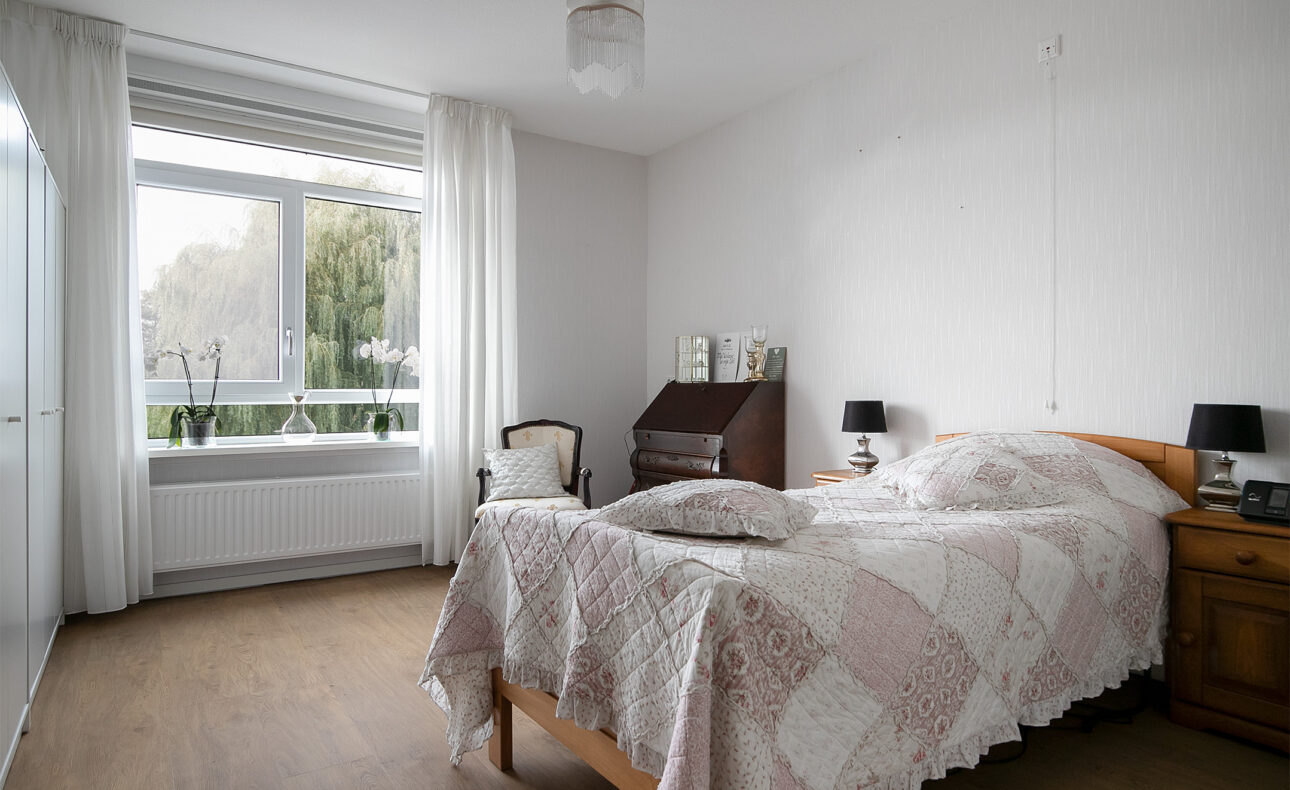 Slaapkamer met een groot raam, een tweepersoonsbed met geblokt dekbedovertrek, houten vloer en een kleine houten kast.
