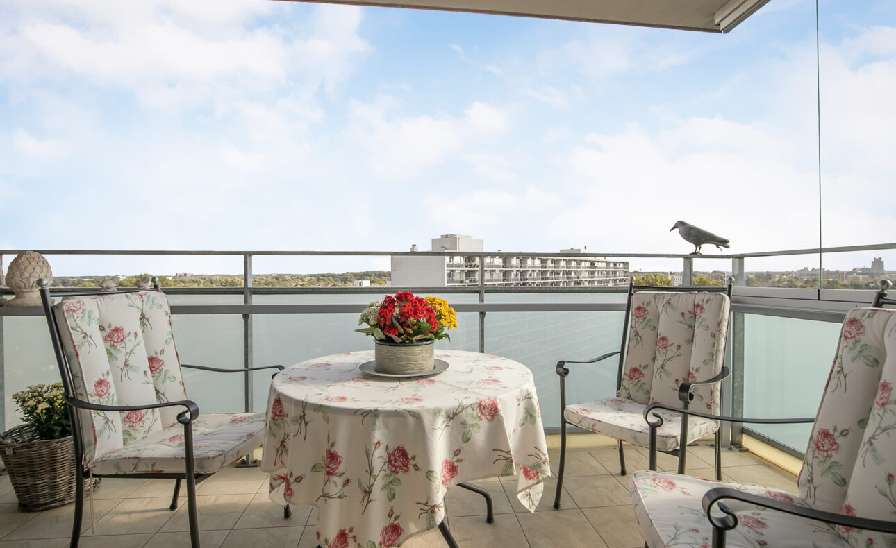 Balkon met uitzicht op water, ronde tafel met bloemen in het midden, omringd door drie stoelen met bloemenpatroon.