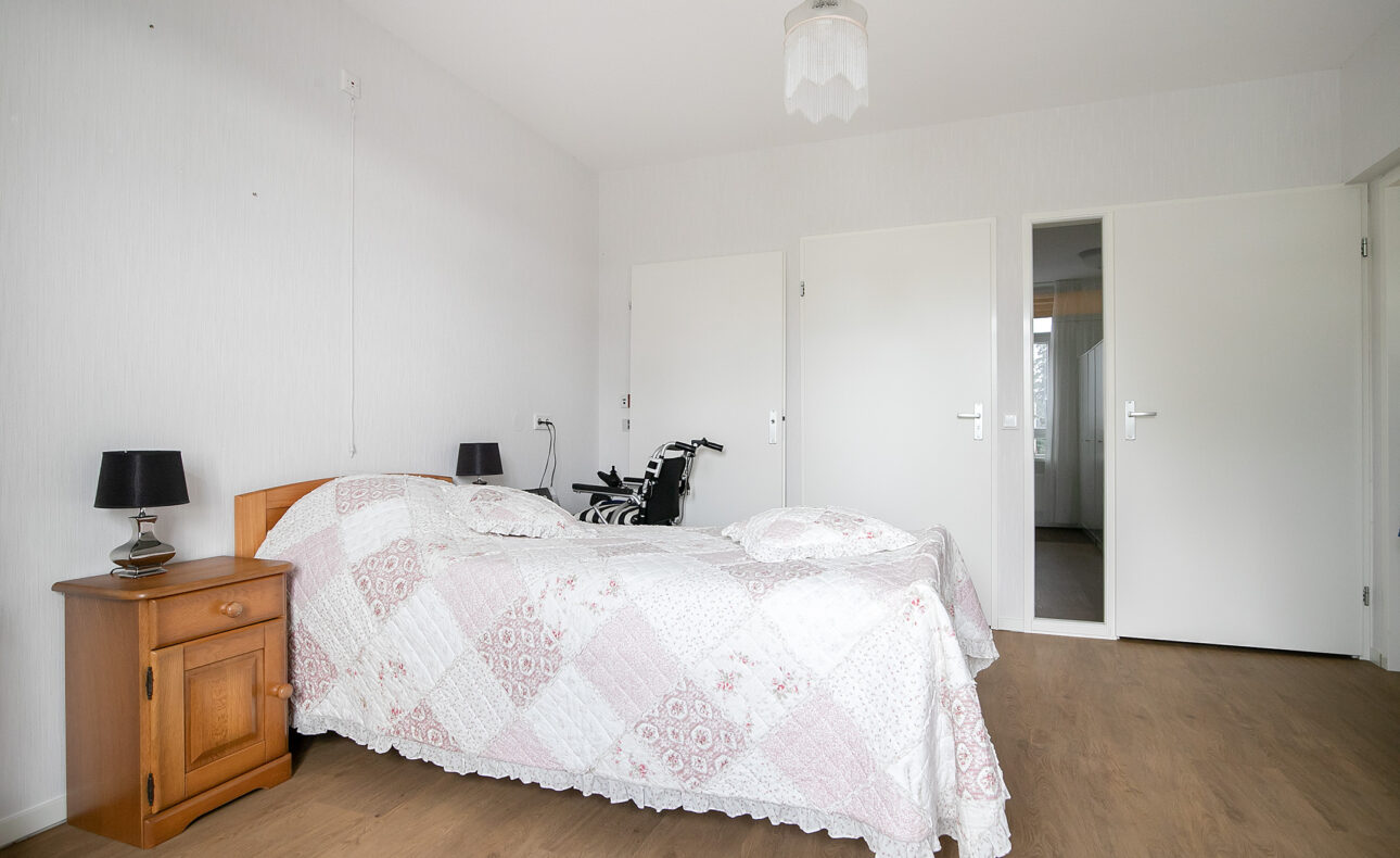Een lichte slaapkamer met een houten bed, wit beddengoed, een nachtkastje met lamp en een rolstoel naast het bed.