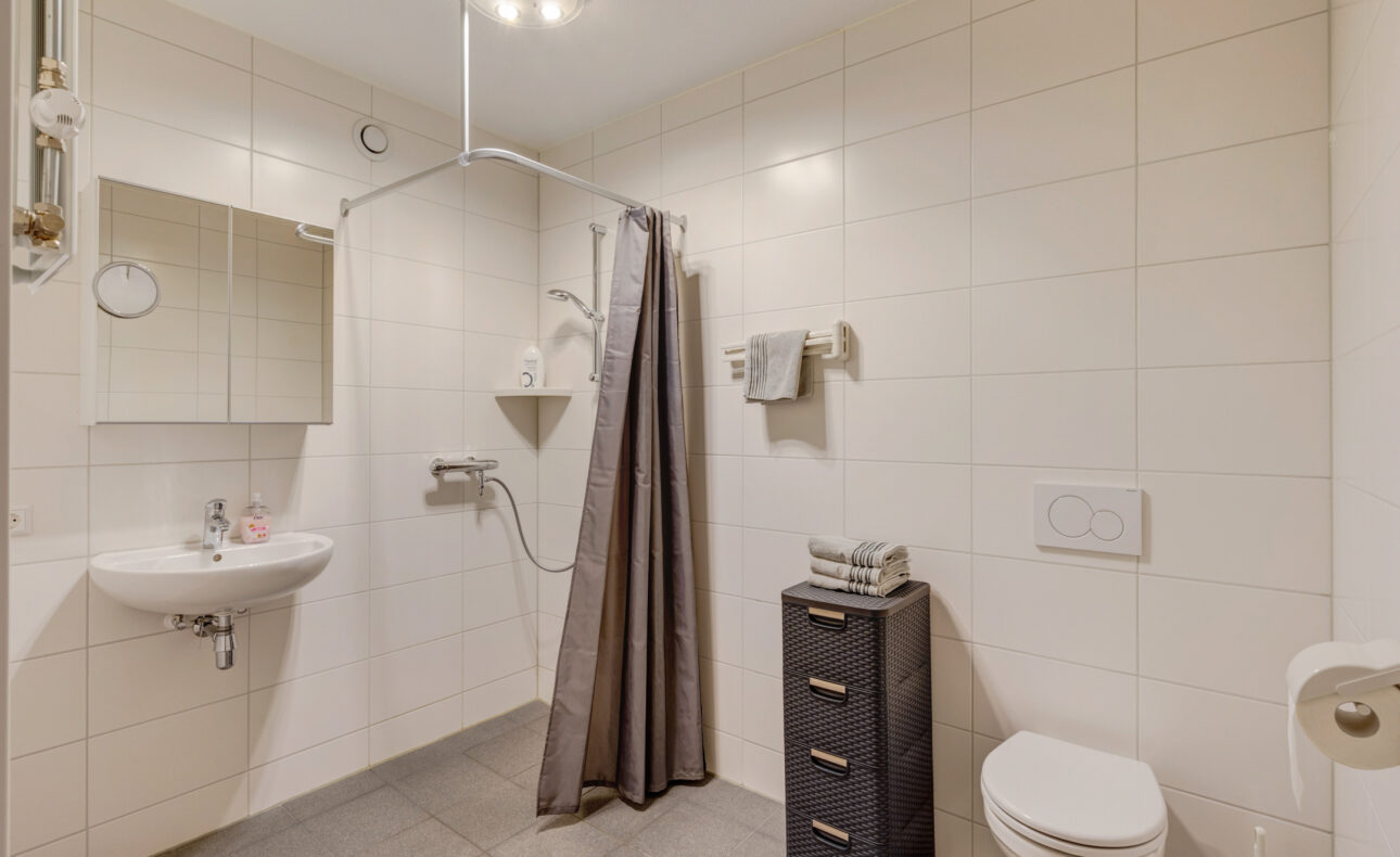 Moderne badkamer met witte tegels, een douche met grijze gordijn, wastafel met spiegel, toilet en kastje met handdoeken.