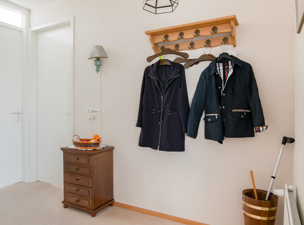 Een hal met een houten kapstok waarop twee jassen hangen, een kleine houten ladekast en een paraplubak met een wandelstok.