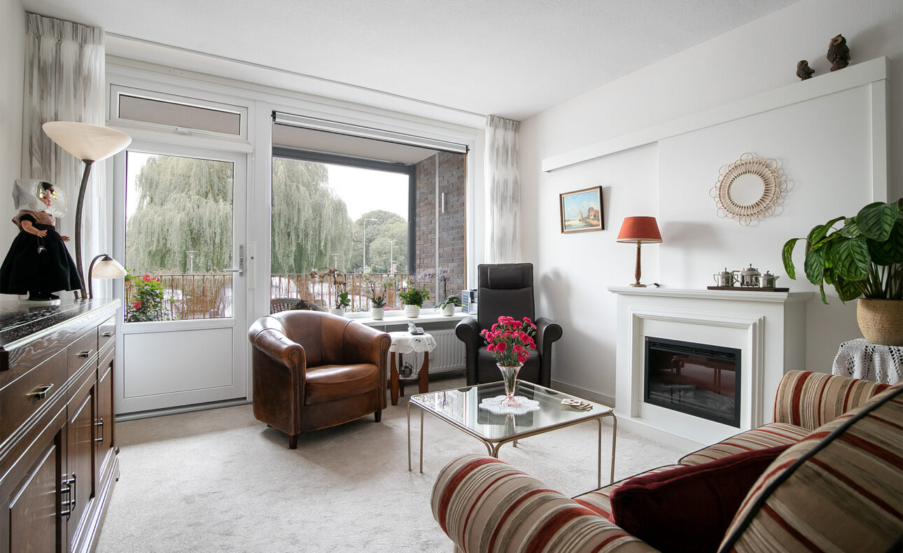 Woonkamer met leren stoel, gestreepte bank, open haard, en glazen salontafel met bloemen. Grote ramen met uitzicht op bomen.