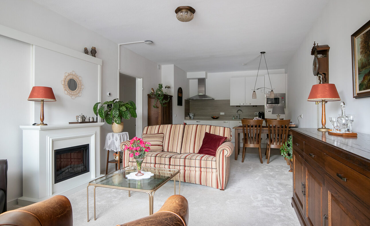 Woonkamer met gestreepte bank, glazen salontafel, open haard, en eettafel. Keuken op de achtergrond. Gezellige sfeer.