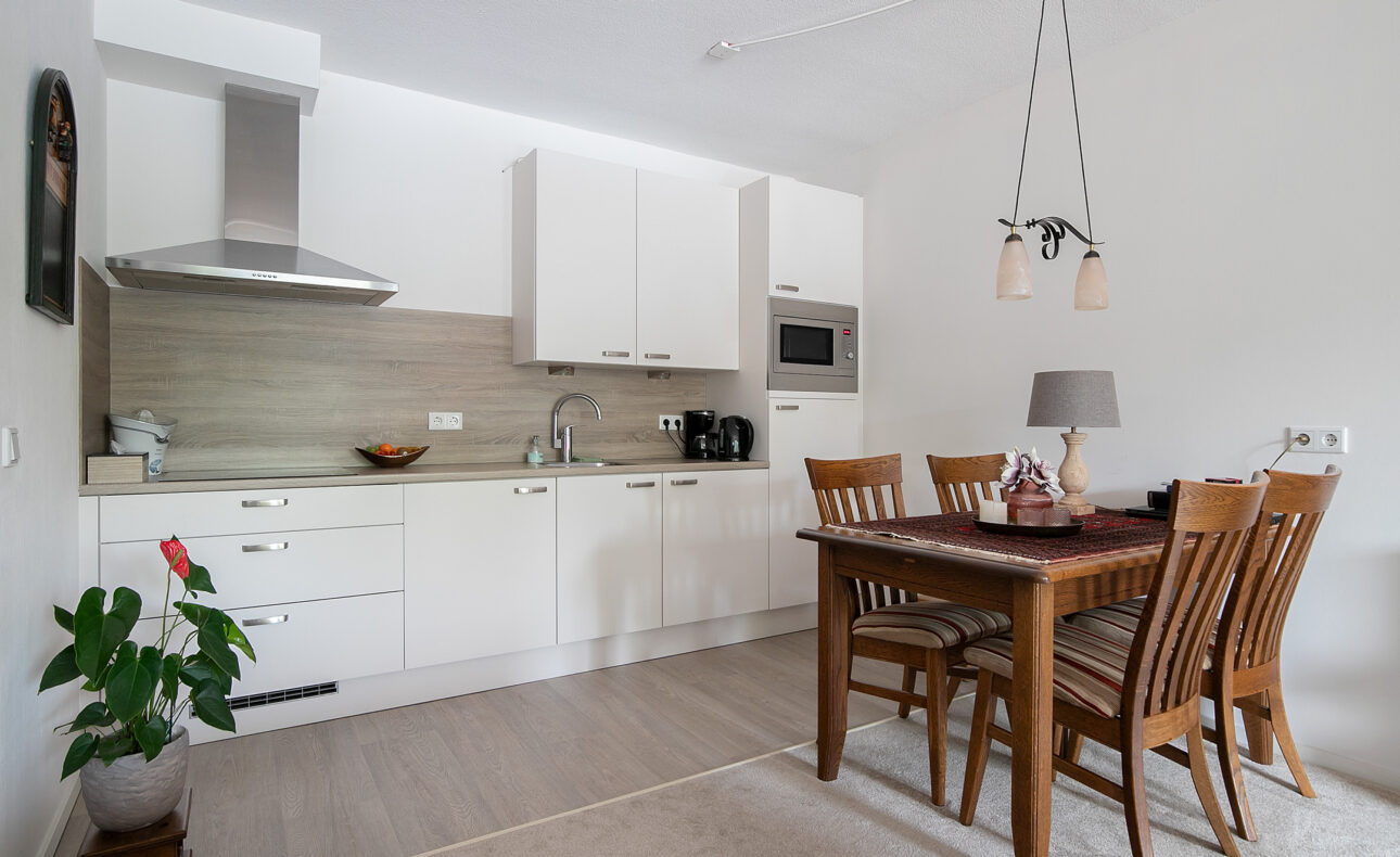 Keuken met witte kasten, houten eettafel en stoelen. Planten en decoraties sieren de ruimte. Moderne en gezellige sfeer.
