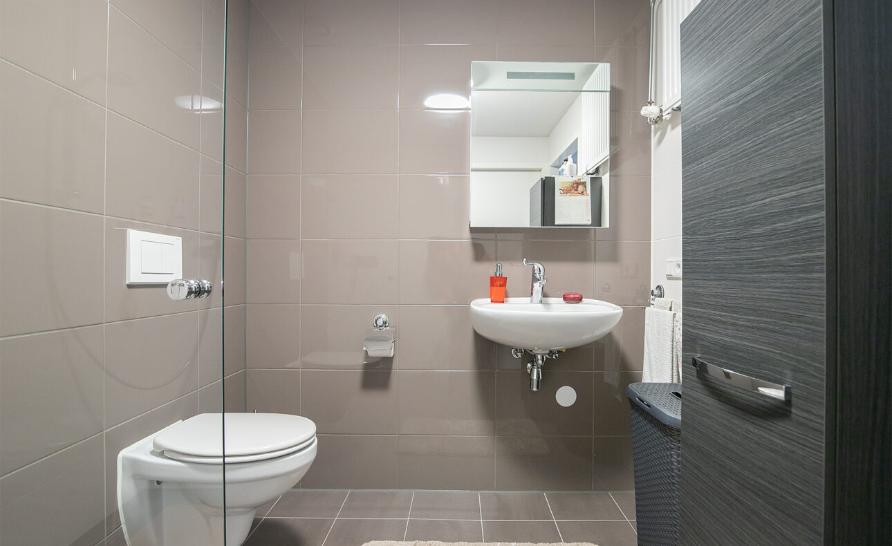 Moderne badkamer met hangend toilet, wastafel met spiegel, glazen douchewand en bruine tegels op vloer en muren.