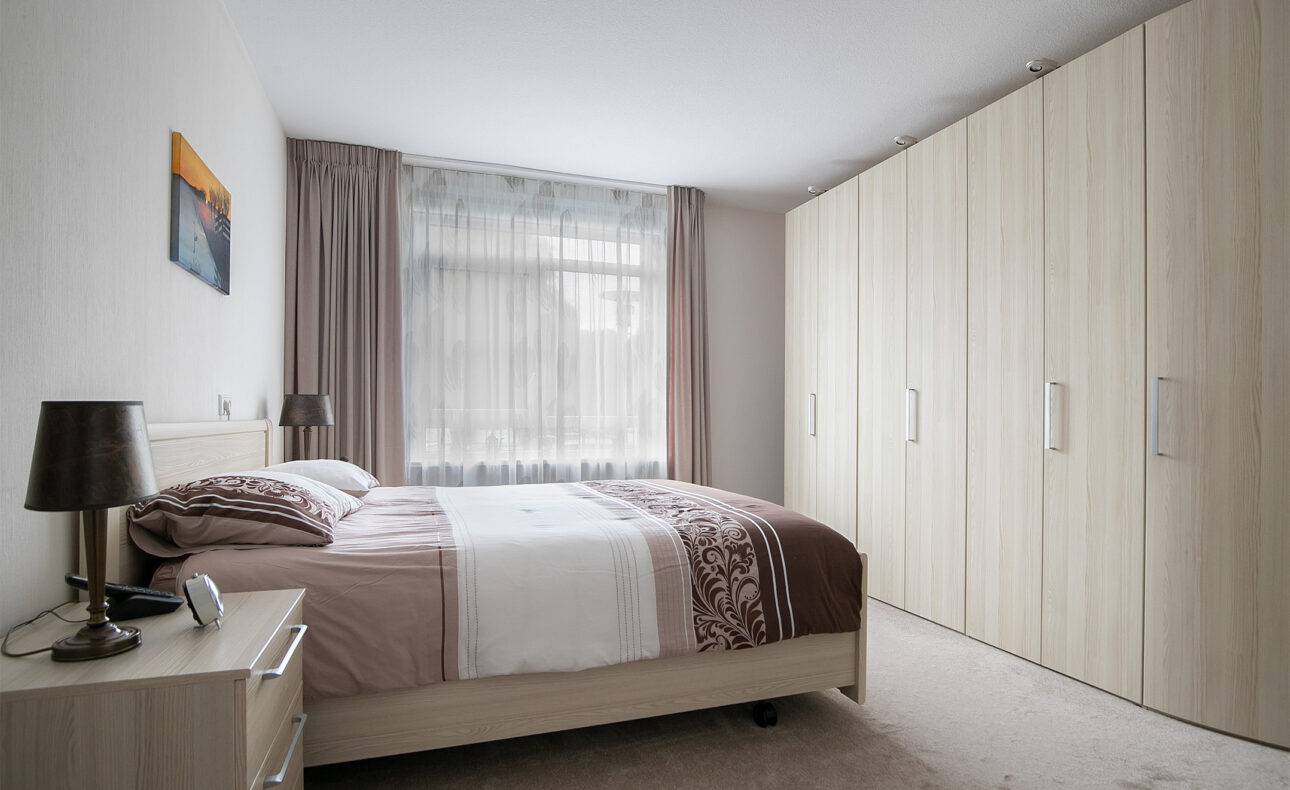Slaapkamer met een tweepersoonsbed, beige dekbed, houten nachtkastje met lamp en een grote kledingkast tegen de muur.