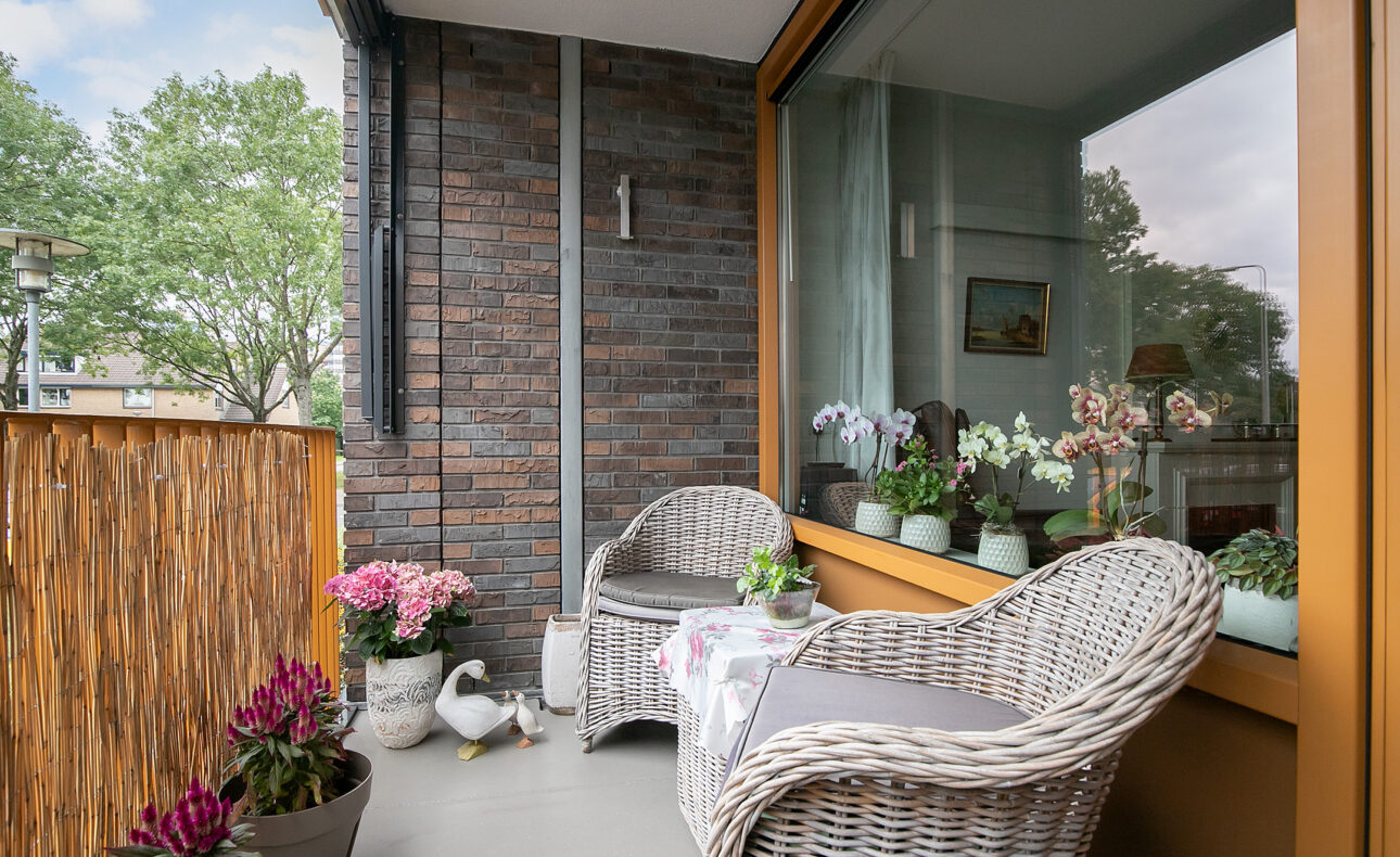 Gezellige balkon met twee rotan stoelen, kleurrijke bloemen en planten, en een eenddecoratie. Bakstenen muur op de achtergrond.