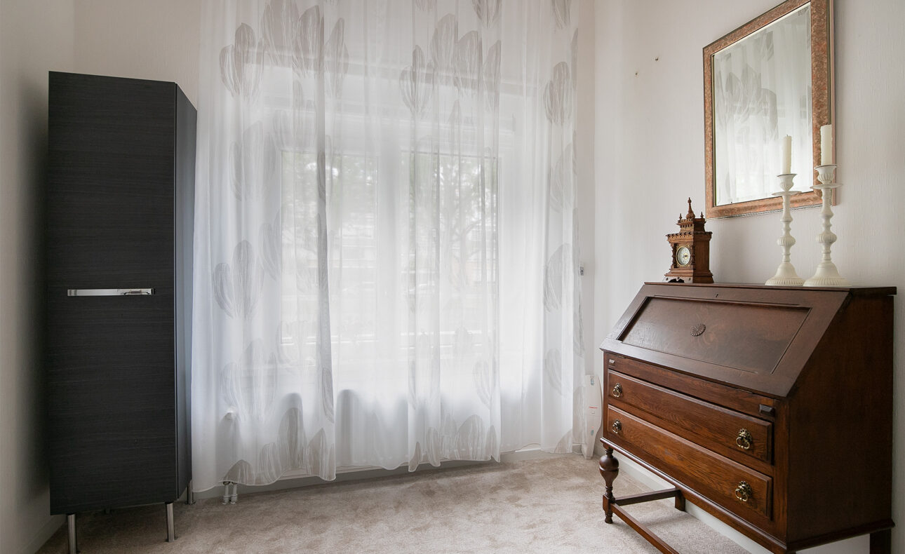 Een lichte kamer met een houten bureau, een hoge kast, een spiegel aan de muur en gordijnen die zachtjes het licht filteren.