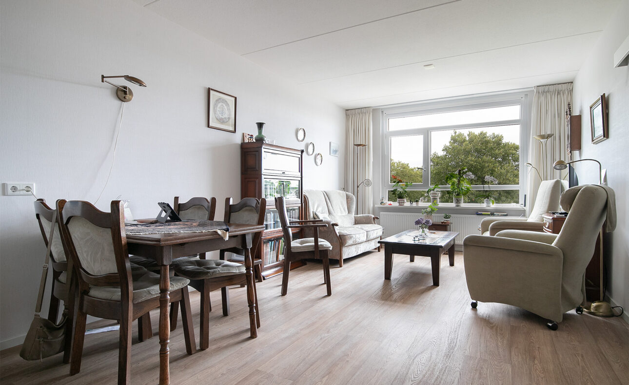 Woonkamer met houten vloer, eettafel met stoelen, comfortabele zitstoelen en een groot raam met uitzicht op groen.