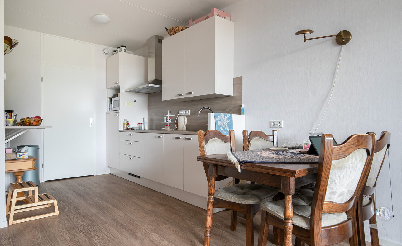 Keuken met witte kasten, houten eettafel en stoelen. Aanrecht met keukengerei, en een wandlamp aan de muur. Houten vloer.