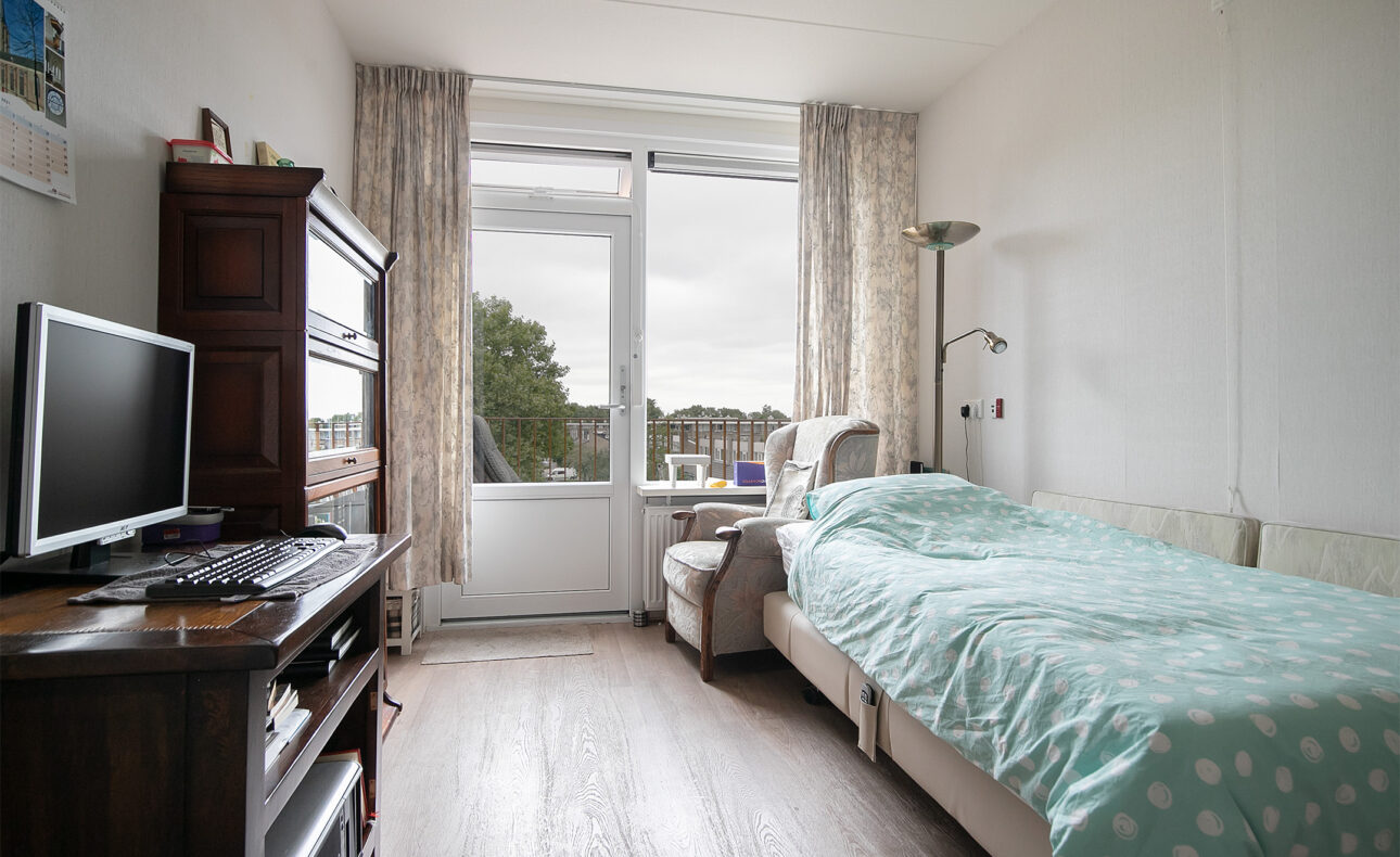 Een lichte kamer met een bed, een stoel, een televisie op een kast en een groot raam met uitzicht op bomen en gebouwen.