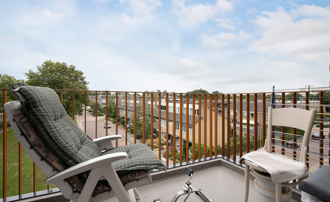 Balkon met twee stoelen, een met kussen, en een rollator. Uitzicht op een groene omgeving en een bewolkte lucht.