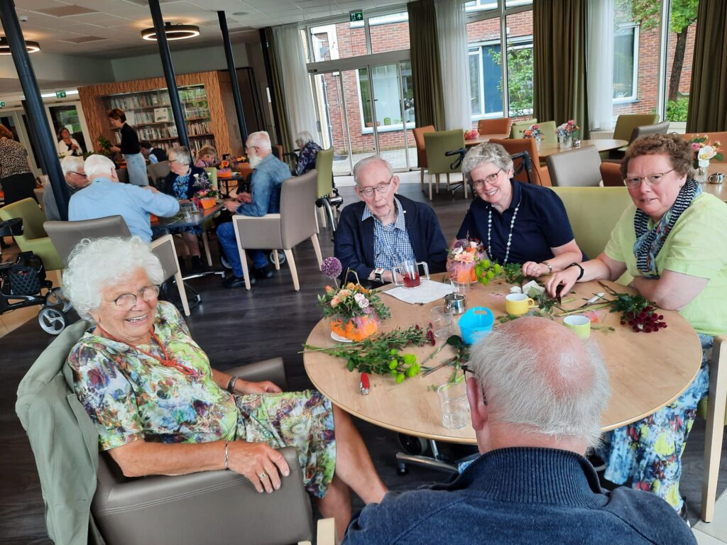 Groep ouderen zit aan een tafel in een gemeenschappelijke ruimte, bezig met bloemschikken. Er heerst een gezellige sfeer.