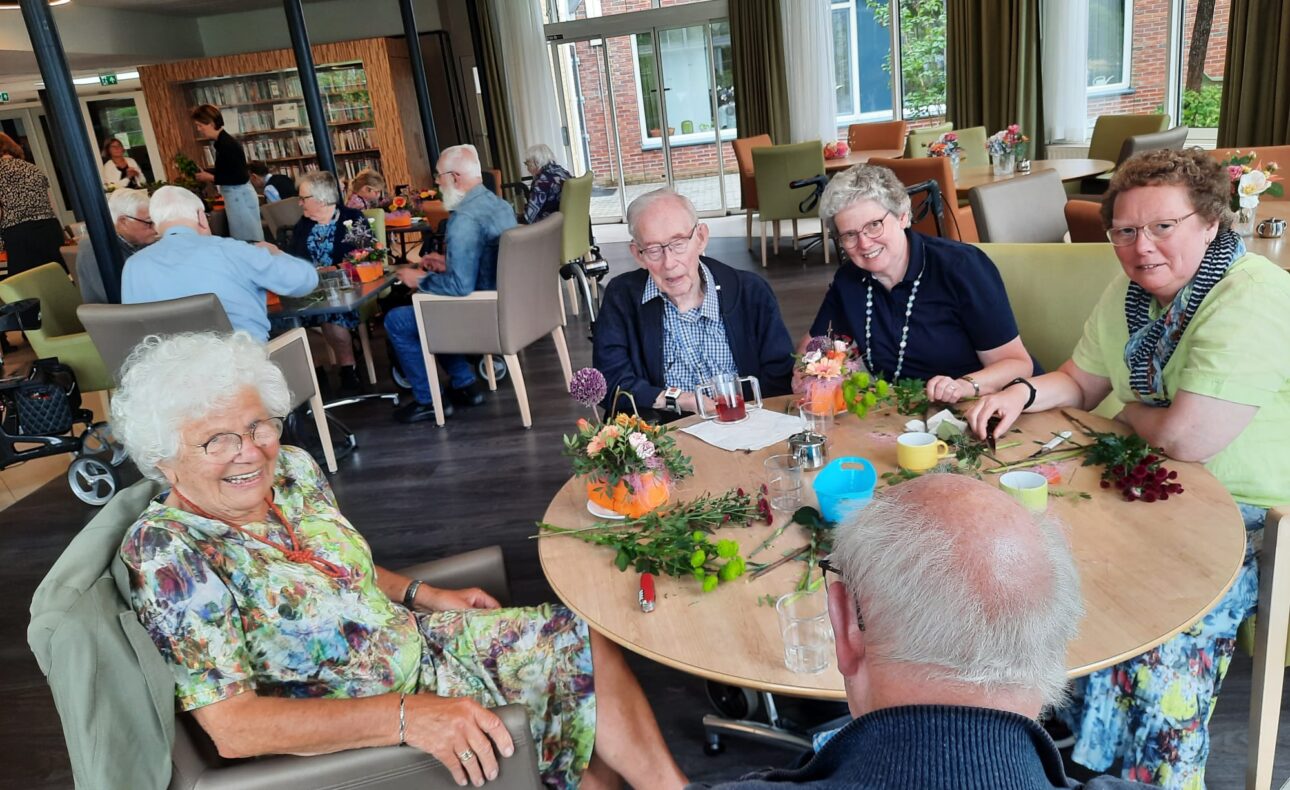 Groep ouderen zit aan een tafel in een gemeenschappelijke ruimte, bezig met bloemschikken. Er heerst een gezellige sfeer.