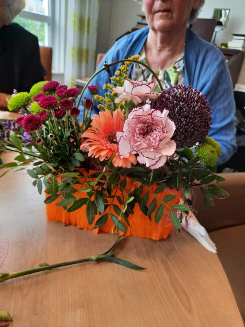 Een kleurrijk bloemstuk met roze, paarse en oranje bloemen in een oranje bakje, op een houten tafel, met een persoon op de achtergrond.