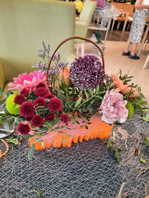 Een kleurrijk bloemstuk in een mandje met paarse, roze en groene bloemen, geplaatst op een tafel met een grijze ondergrond.