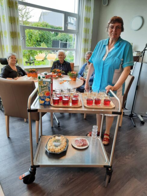 Een vrouw in een blauwe blouse serveert drankjes op een karretje aan een groep ouderen die aan een tafel zitten in een kamer.